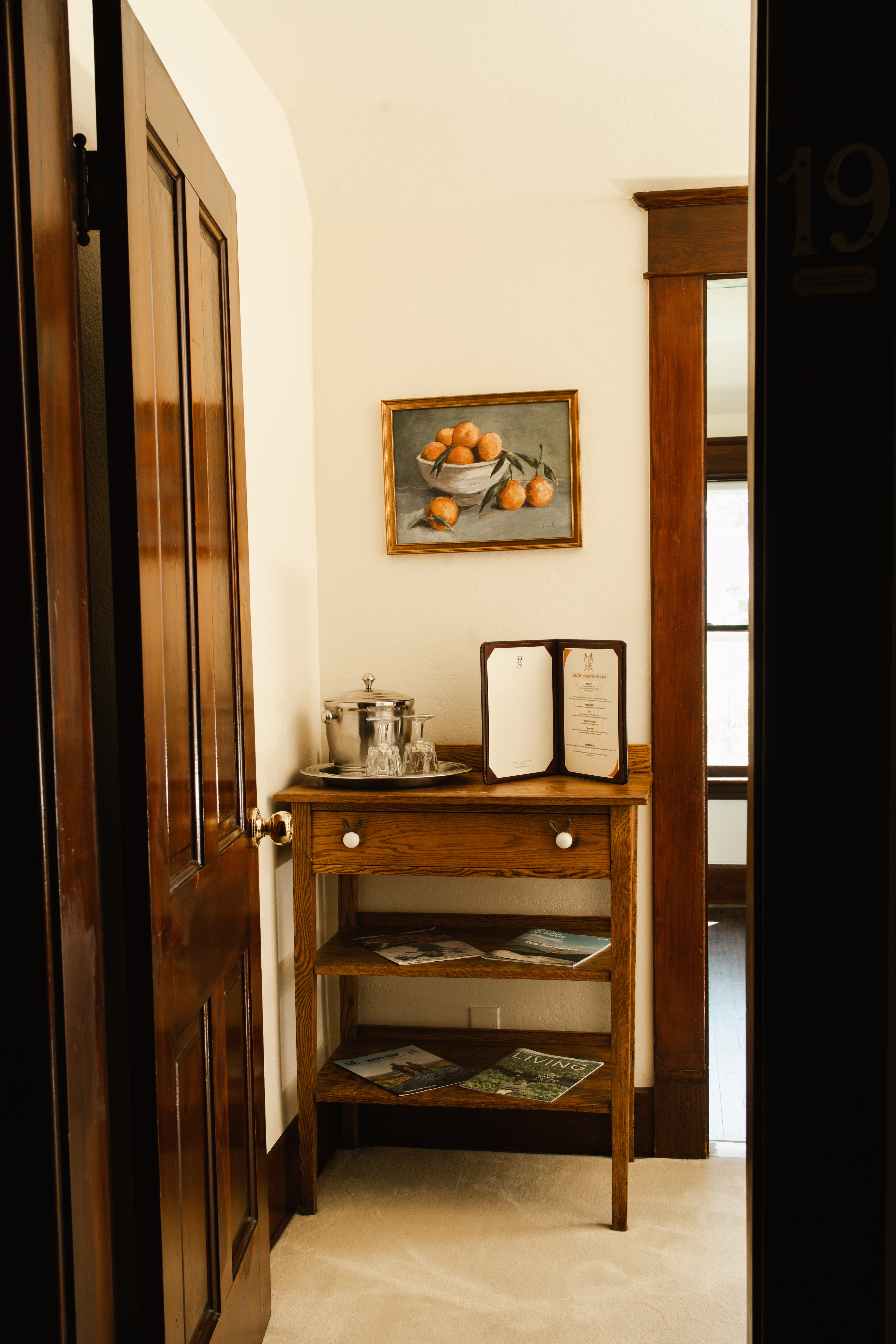 A door opens to a cozy corner with a wooden table displaying a silver tea set, a menu, and magazines, alongside a framed painting of oranges on the wall.