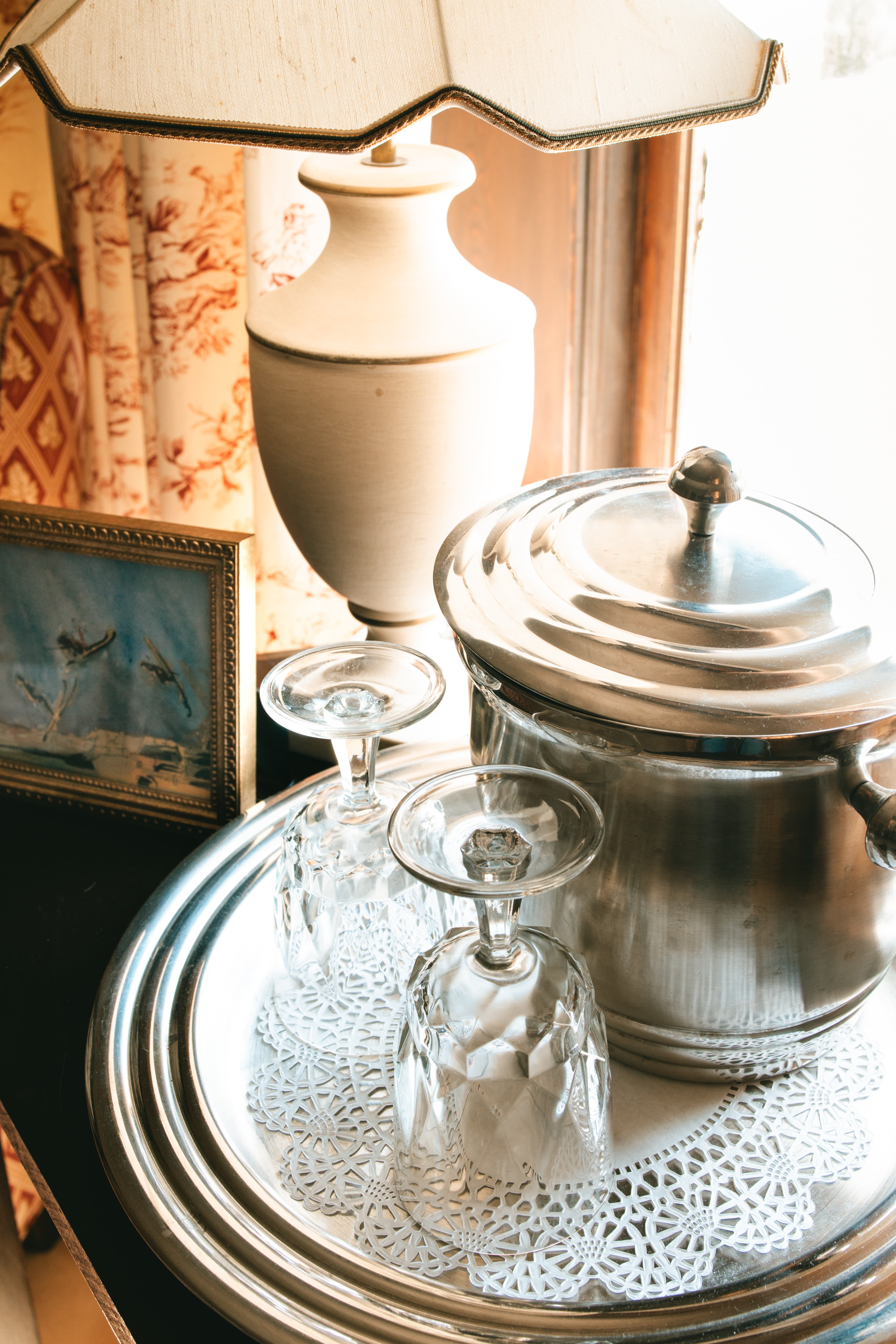 A silver tray with crystal glasses, a metal ice bucket, a lamp, and a framed painting on a wooden table by a window.