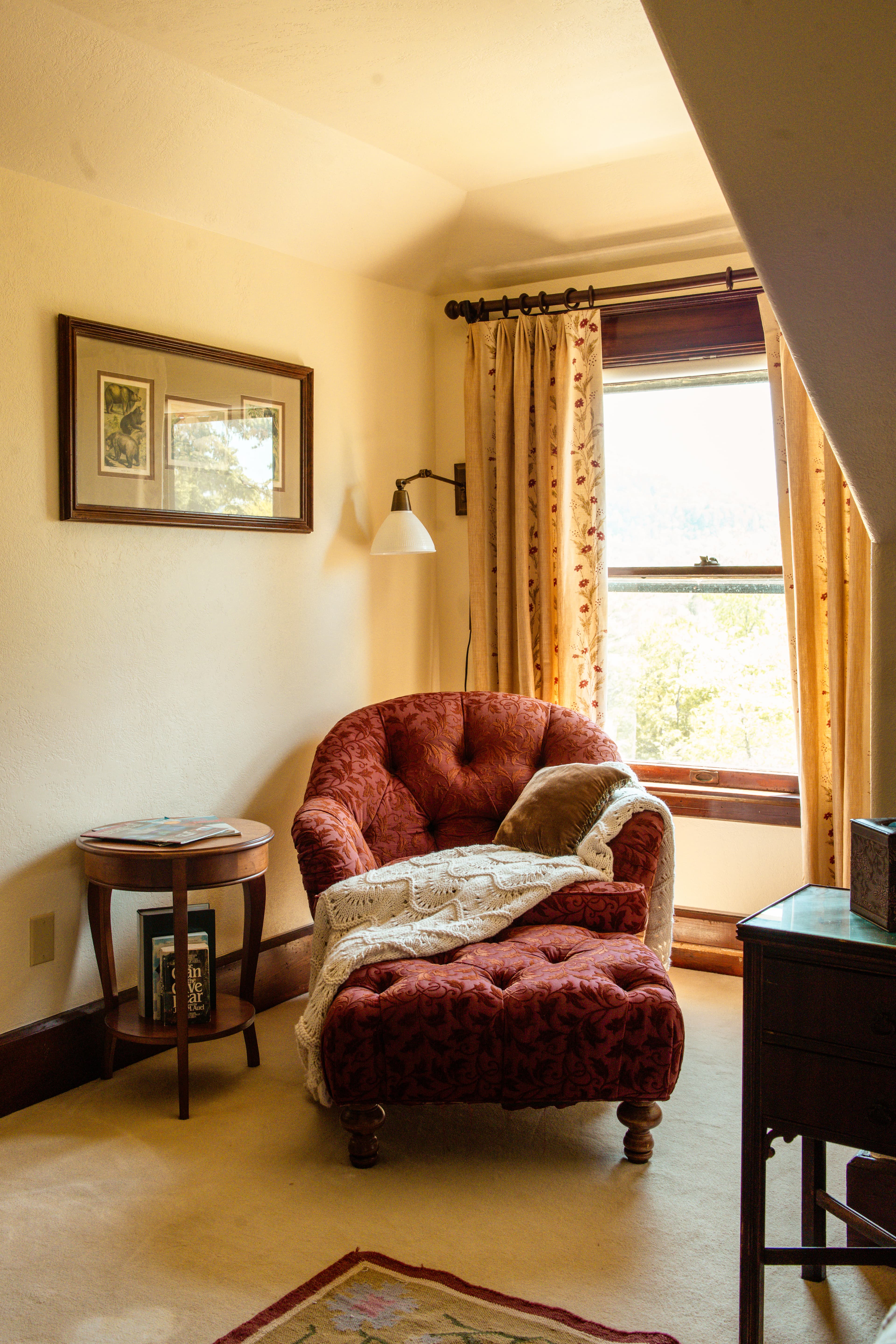 Cozy reading nook featuring a red, tufted chair with a blanket, accompanied by a side table and a window with floral curtains.