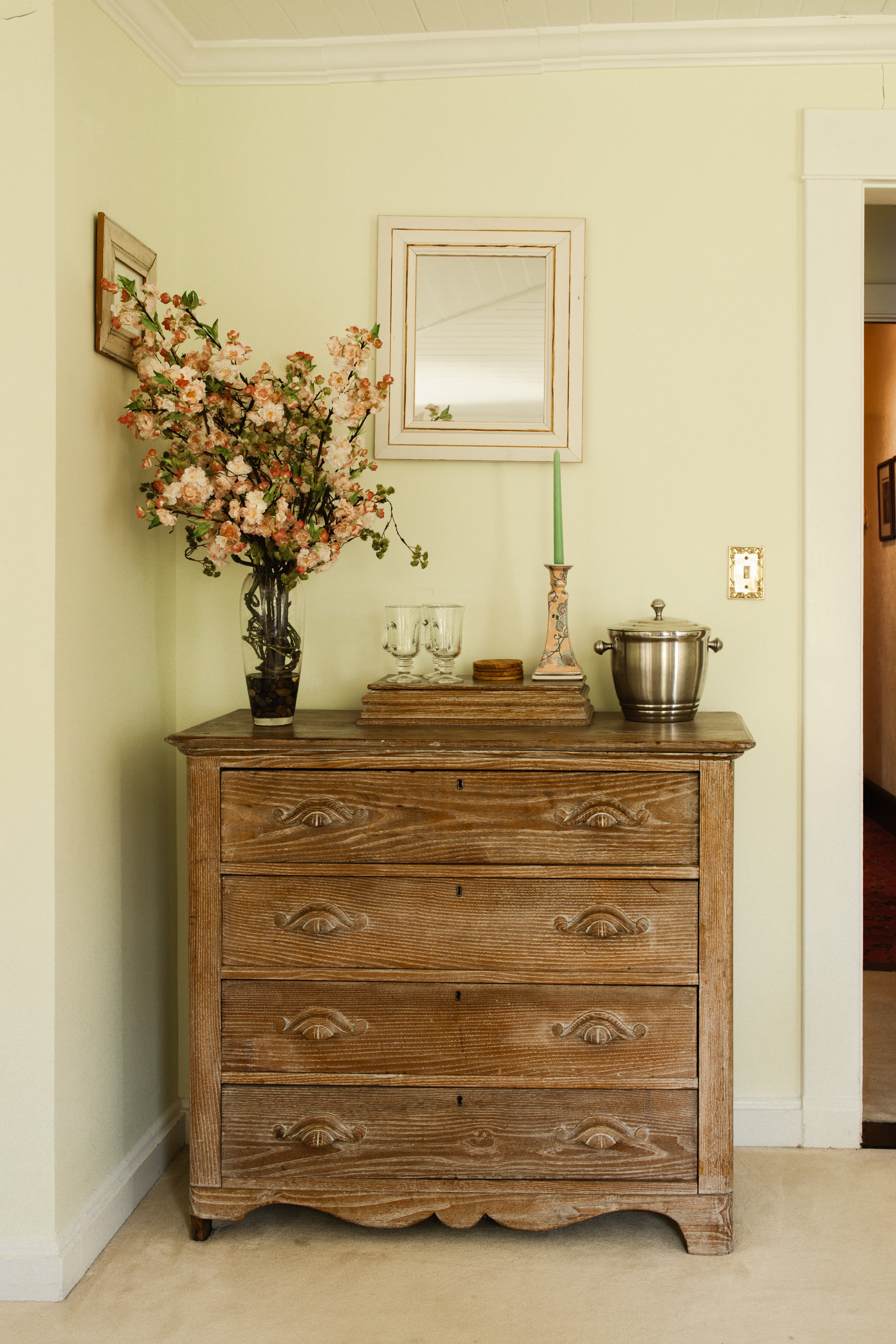 A wooden dresser topped with a vase of flowers, a mirror, a green candle, and a silver ice bucket against a pale green wall.