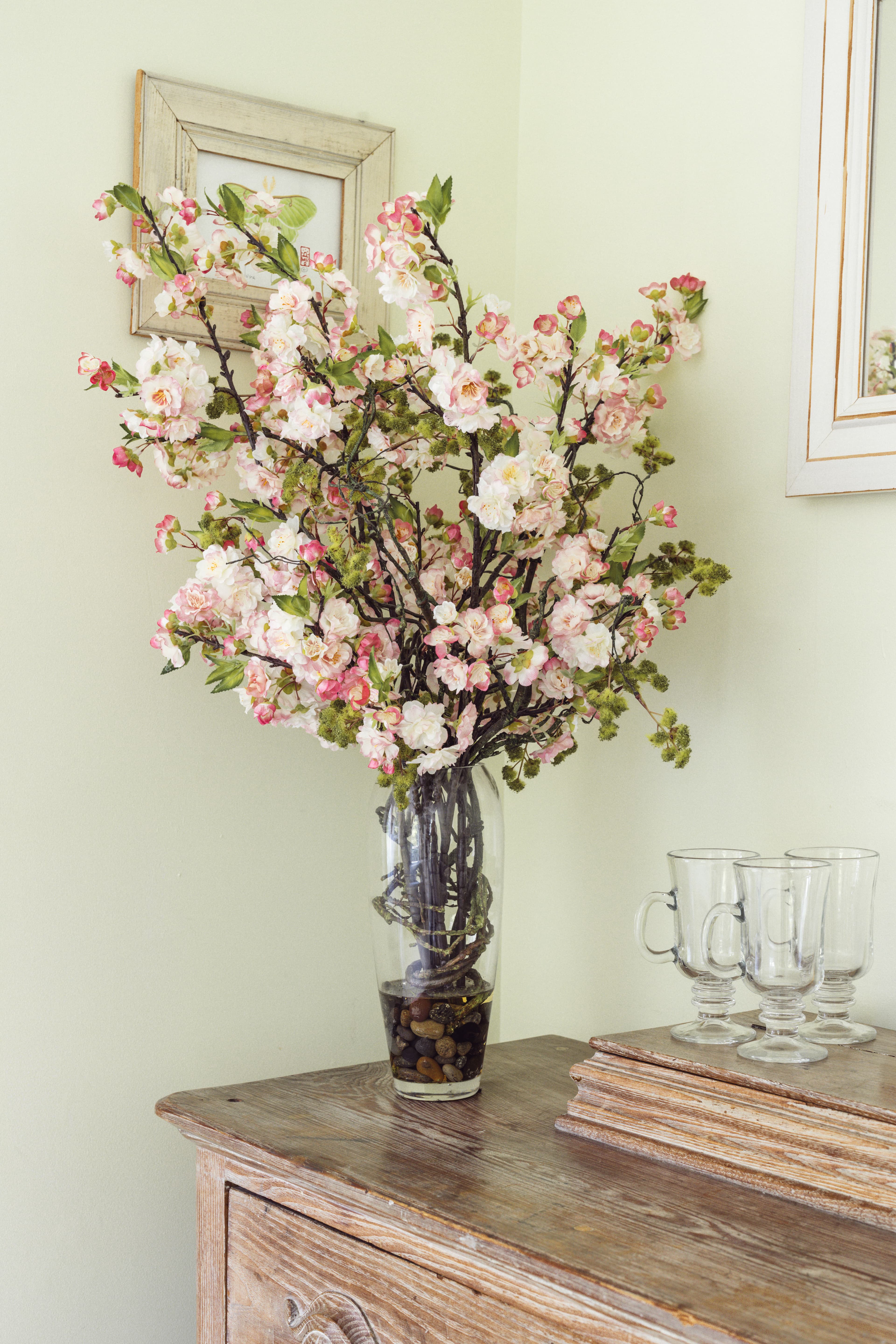 A glass vase filled with pink and white floral arrangements sits on a wooden table next to glassware and a framed picture on a light green wall.