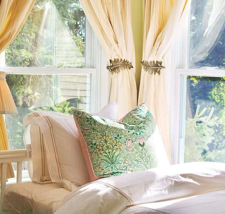 A cozy bedroom corner featuring a decorative pillow and softly flowing curtains by a bright window.