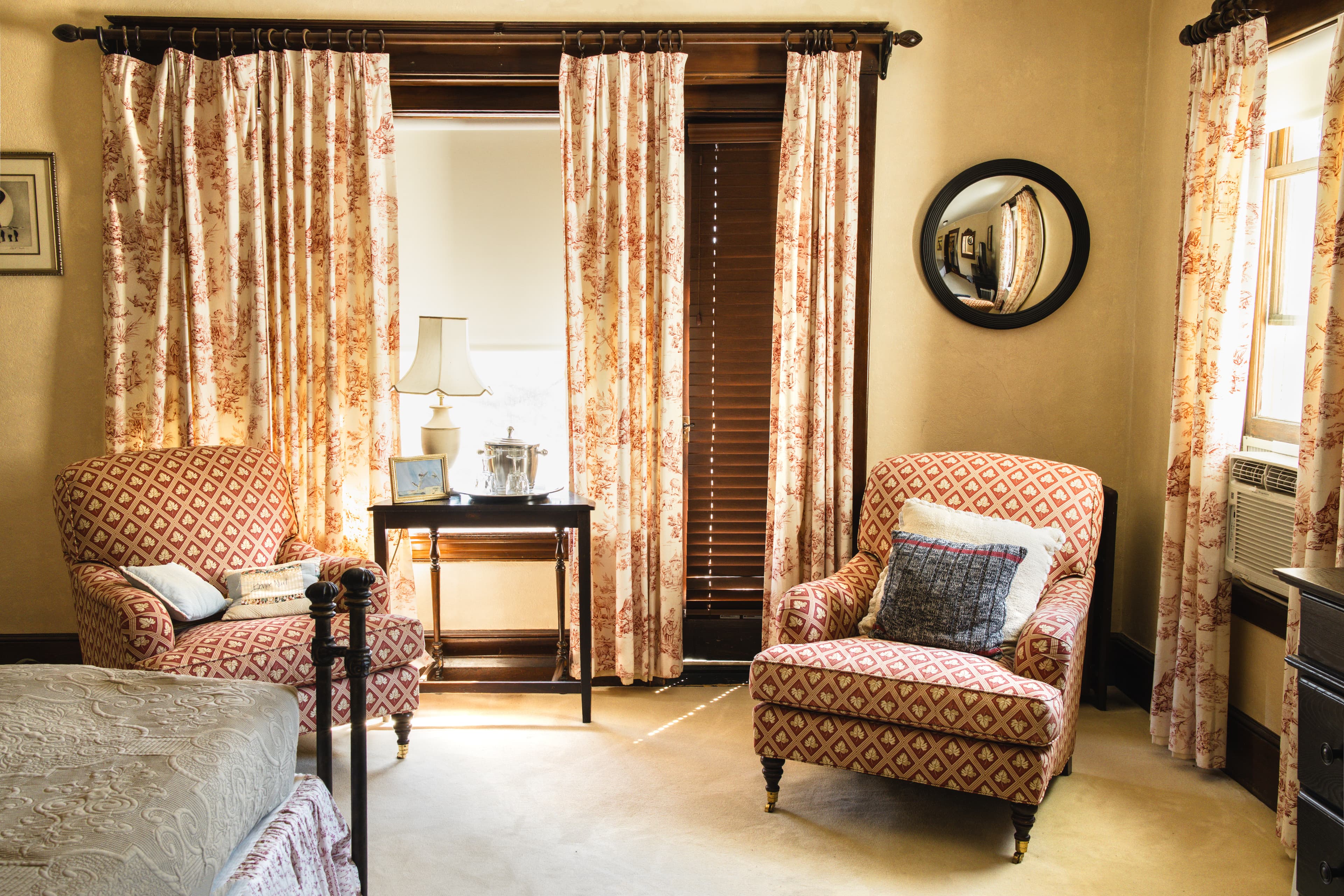 A cozy bedroom corner with patterned armchairs, a small table, and floral curtains against a warm wall.