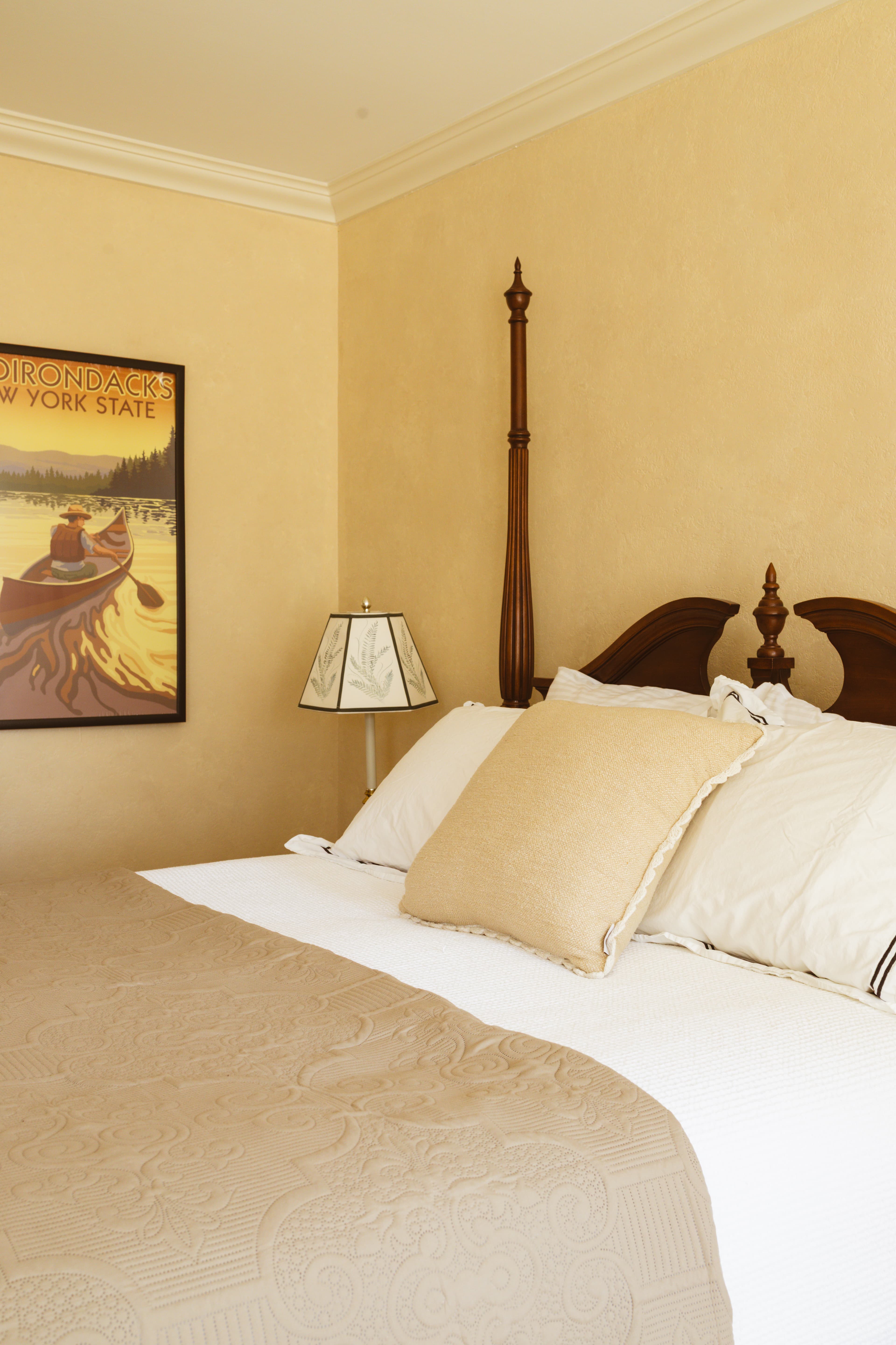A neatly made bed with a decorative quilt and pillows, beside a lamp and a vintage poster on the wall.