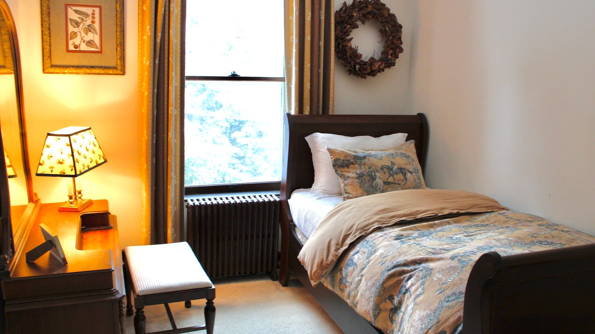 A cozy bedroom featuring a single bed, a wooden dresser, a small bench, and a window with curtains.