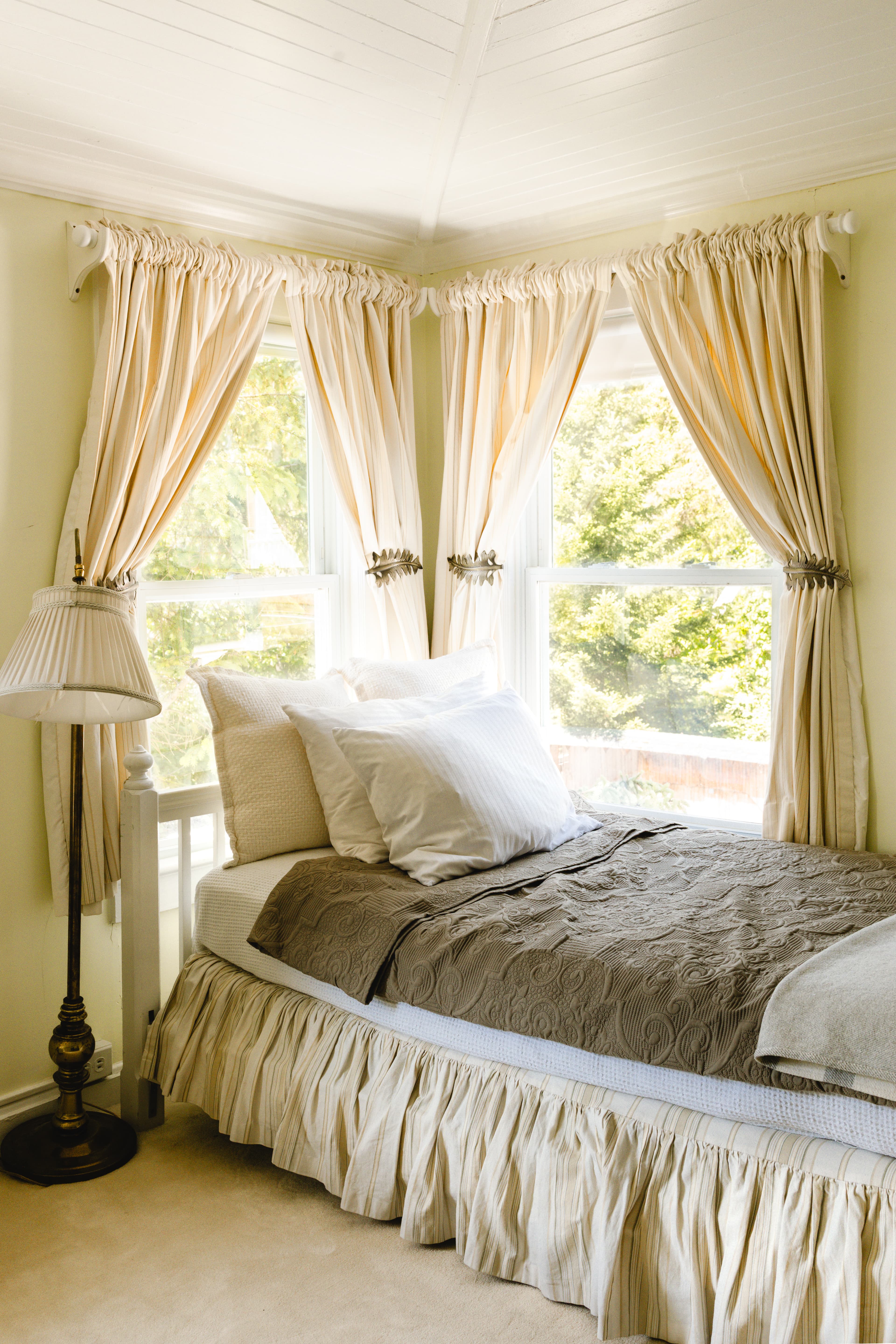 Cozy bedroom with a single bed, decorative pillows, a gray bedspread, and soft curtains beside large windows.