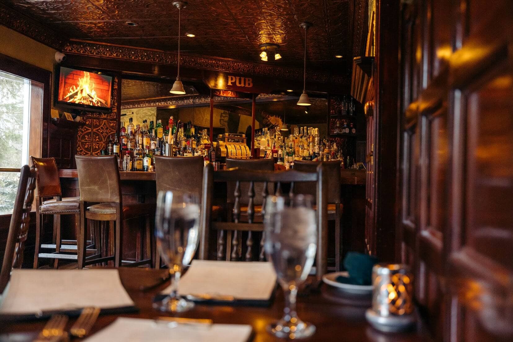 Interior of a cozy pub featuring a wooden bar, shelves of liquor, and dining tables set with water glasses.