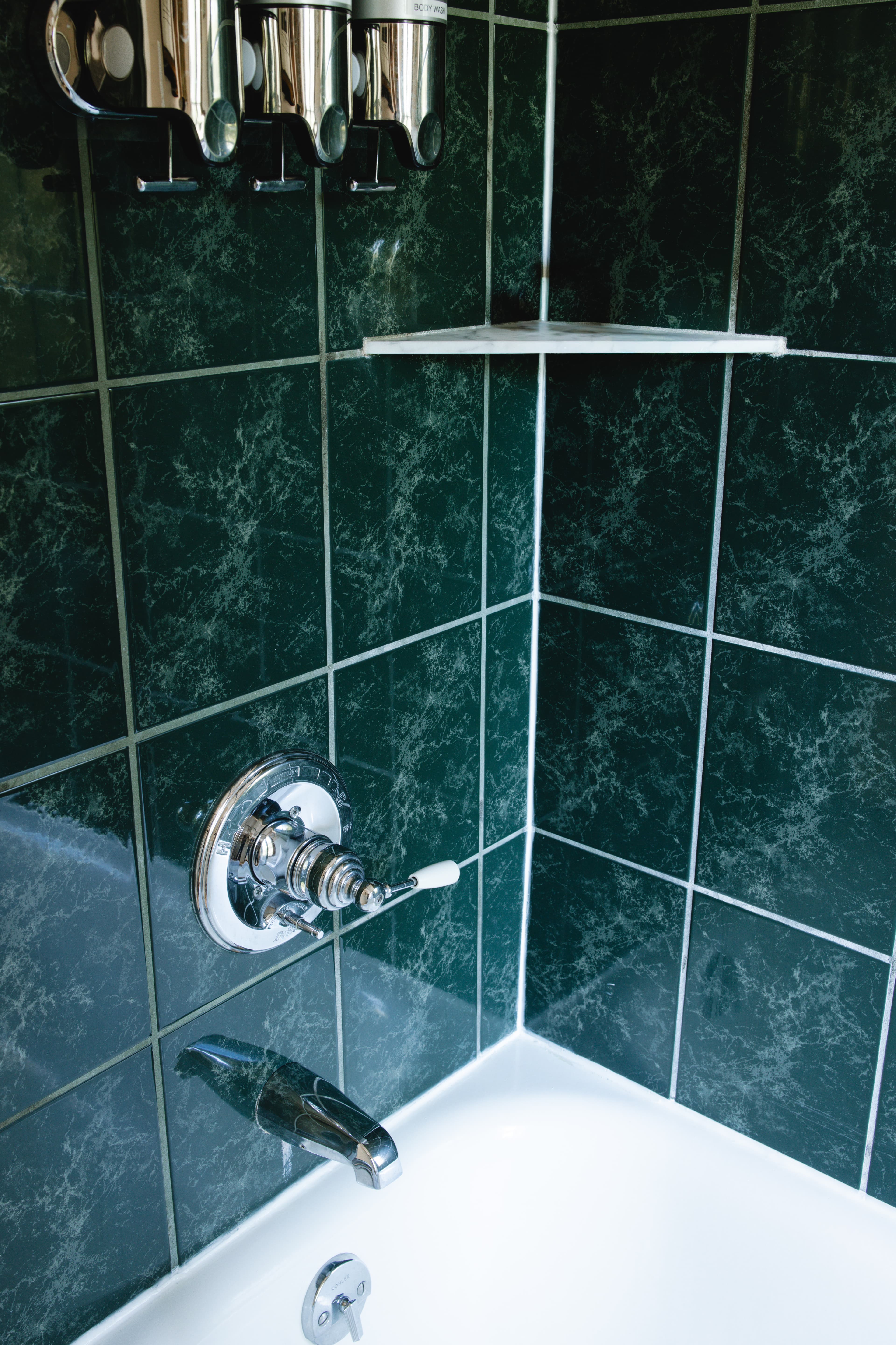 A corner shower featuring green tiled walls, a chrome faucet, and a soap dispenser.