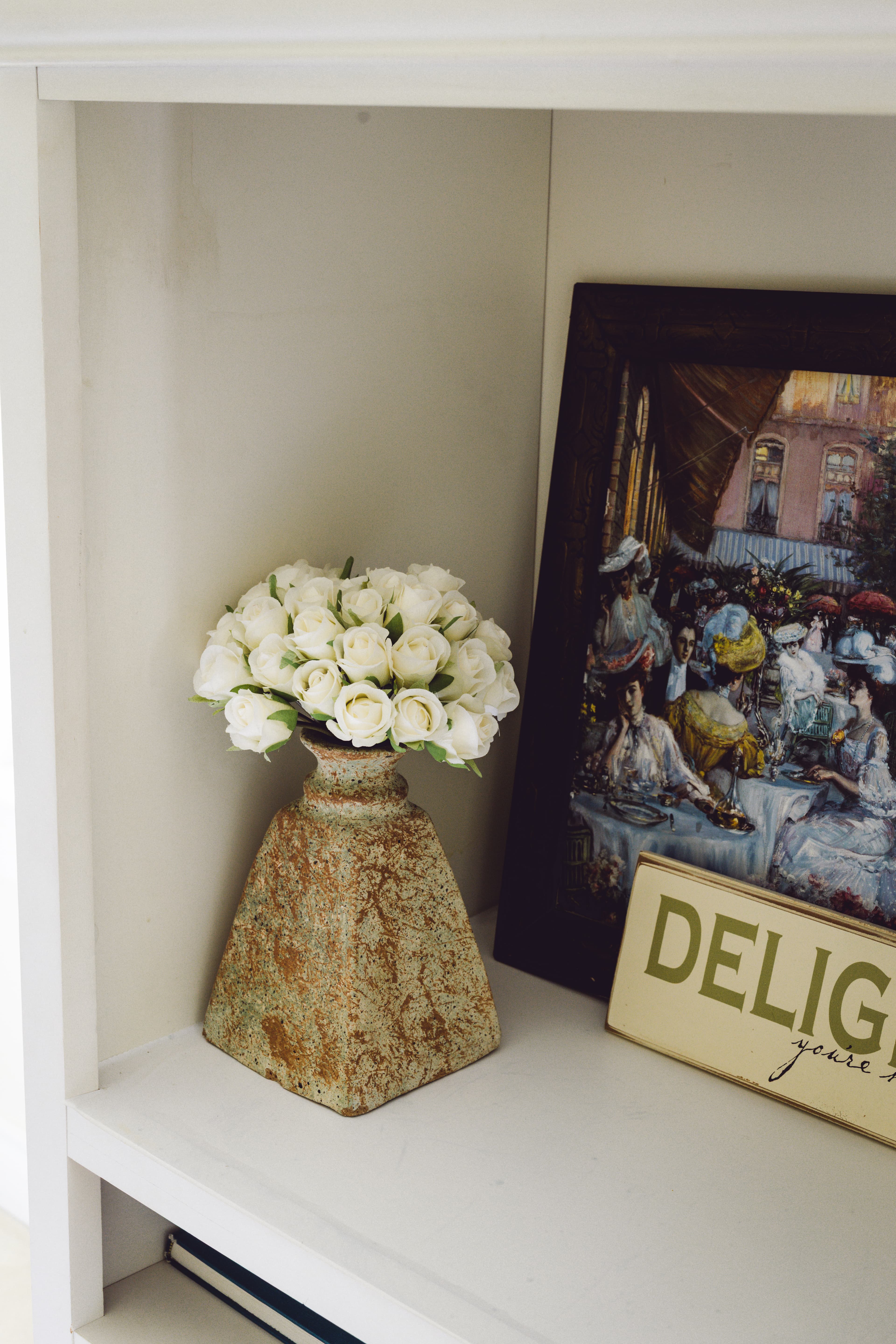 A rustic vase holds white flowers next to a framed artwork and a decorative sign.