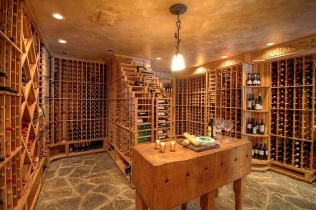 A spacious wine cellar with wooden racks, a stone floor, and a central wooden table holding wine glasses and accessories.