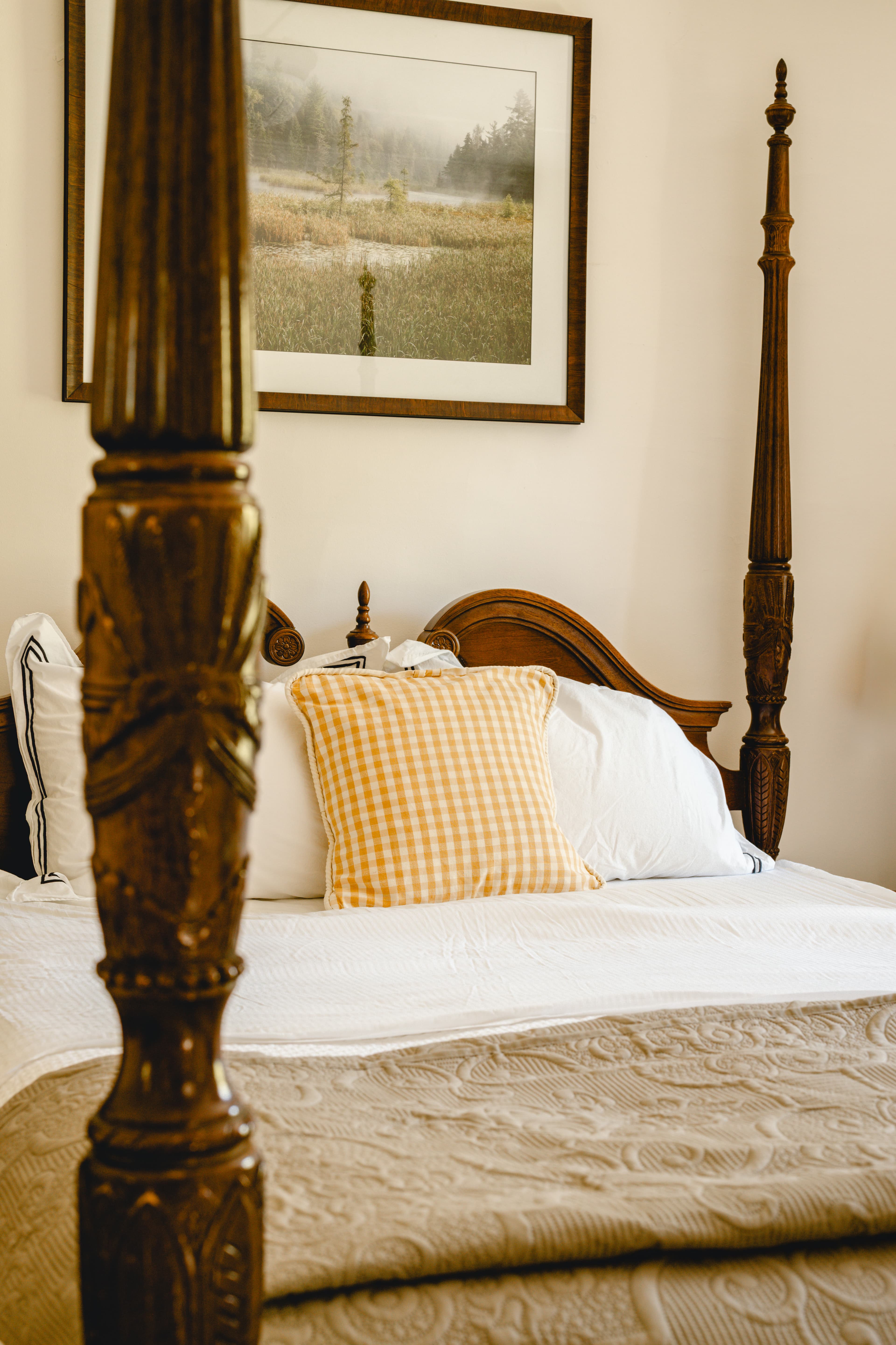 A cozy bedroom featuring a four-poster bed with a gingham pillow and a landscape photograph on the wall.
