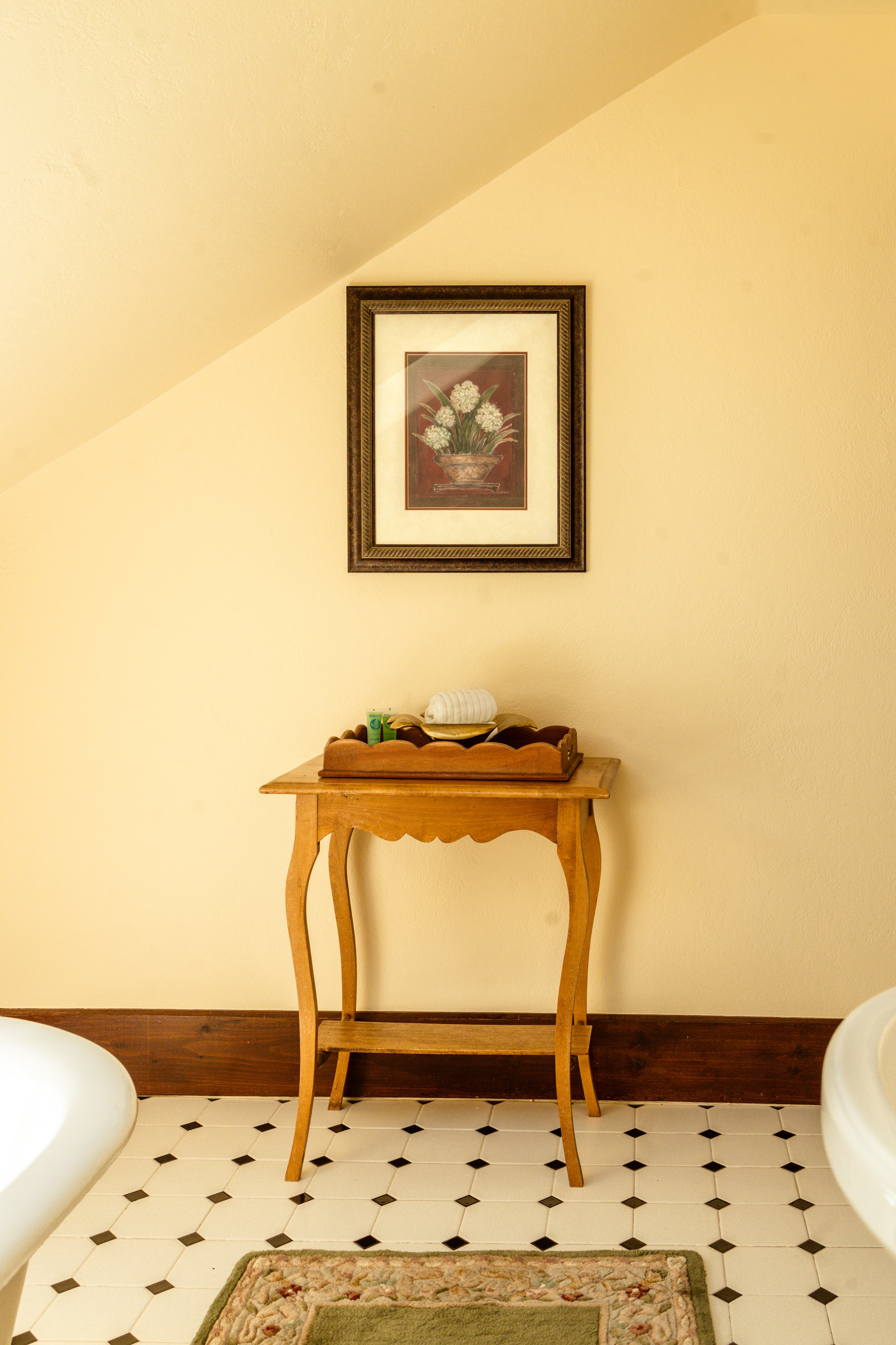 A wooden side table with a decorative tray sits beneath a framed floral picture in a softly lit room.