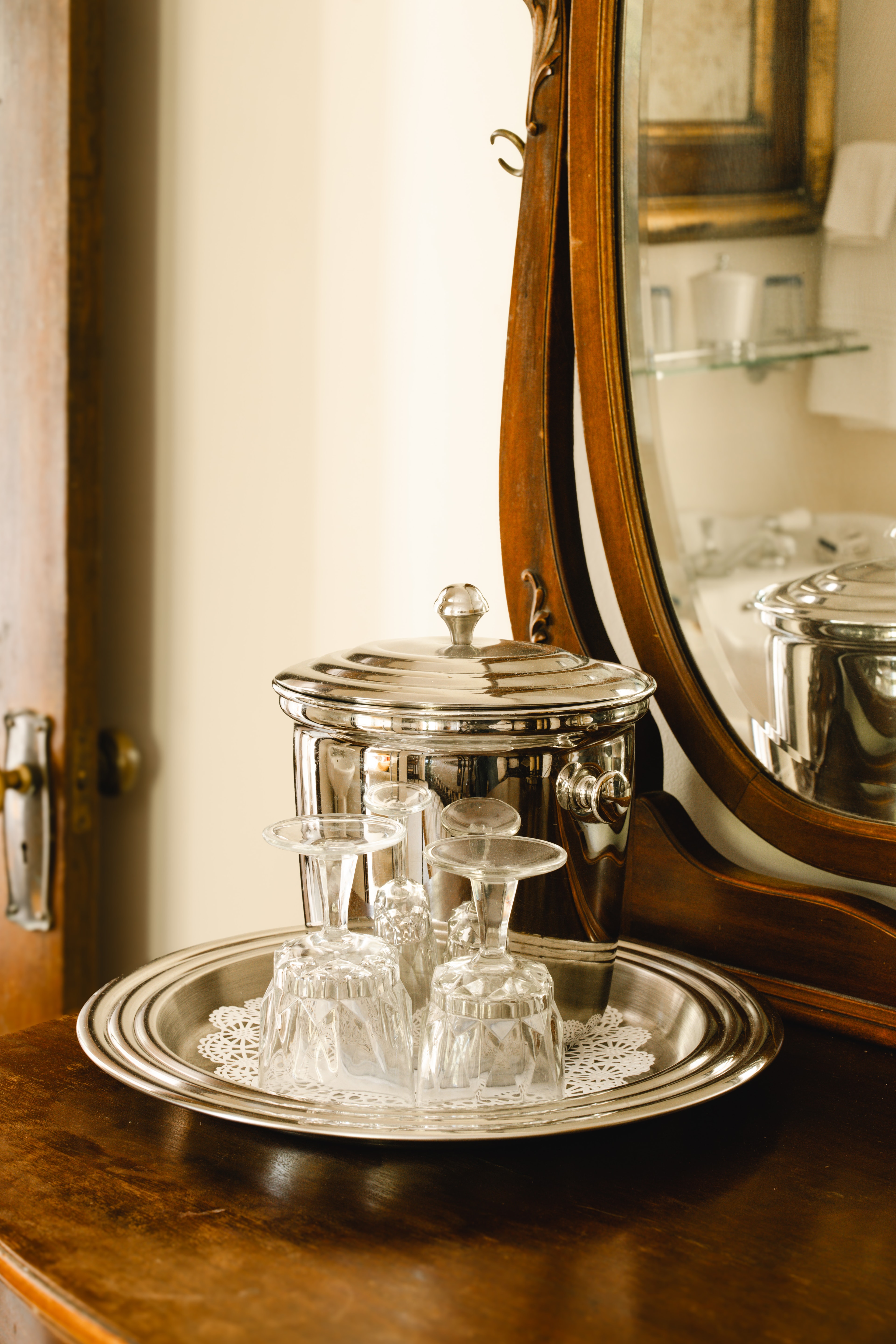 A silver tray with a covered container and several elegant glassware pieces, reflected in a wooden mirror.