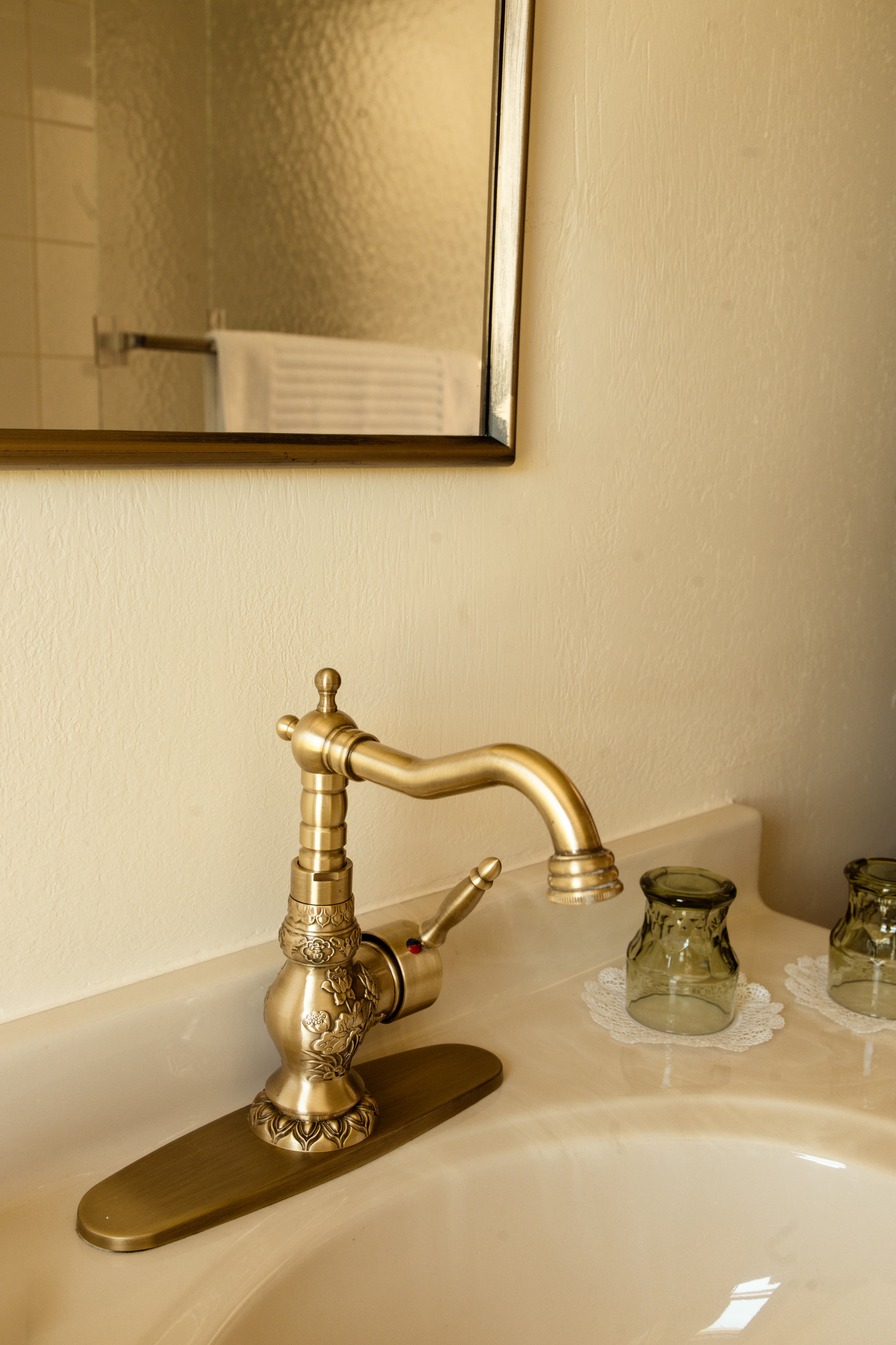 A vintage brass faucet is mounted on a cream-colored sink, next to two glass jars and a decorative lace doily.
