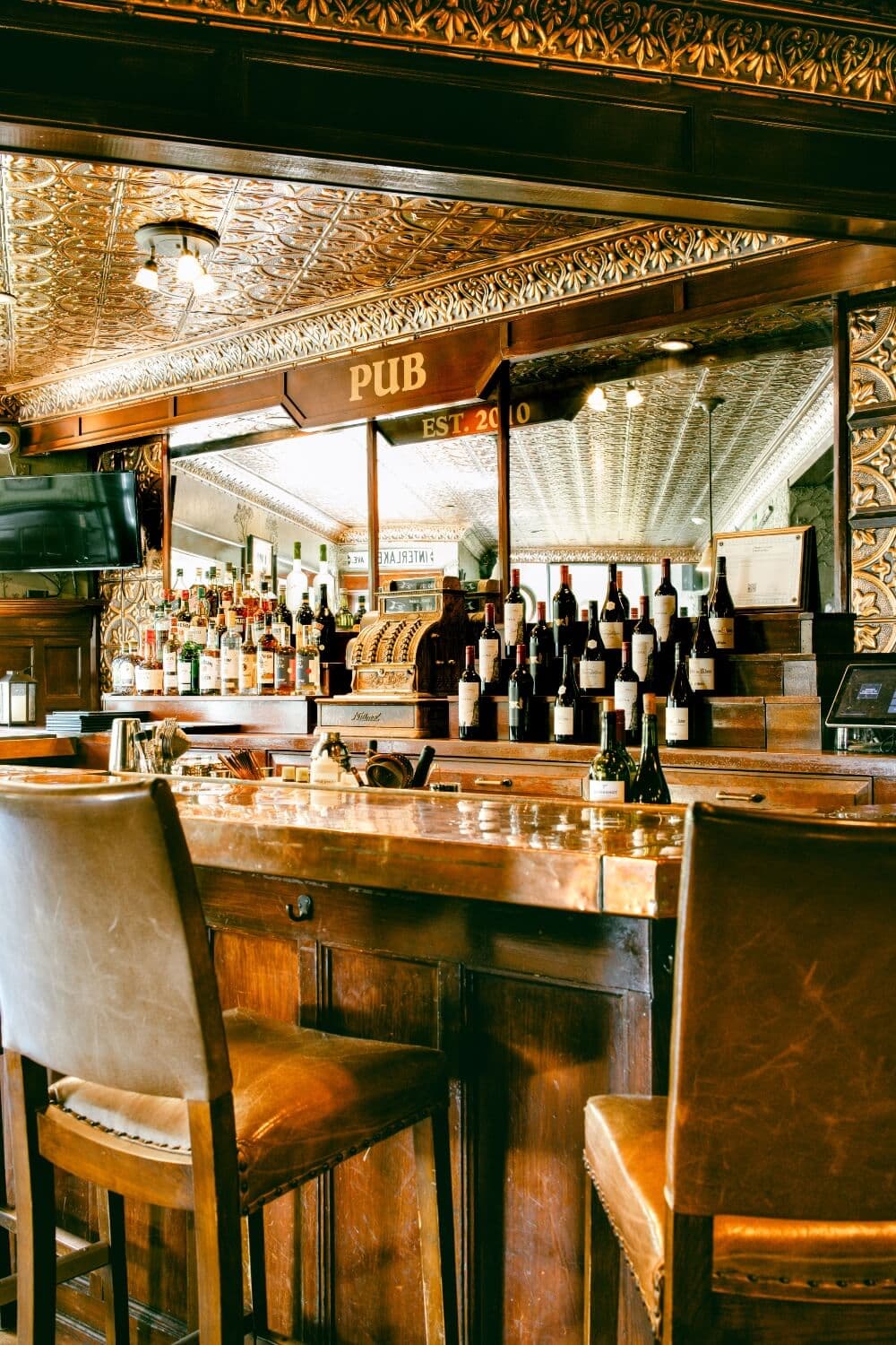 A polished wooden bar in a cozy pub, featuring shelves lined with various bottles of alcohol and a decorative ceiling.