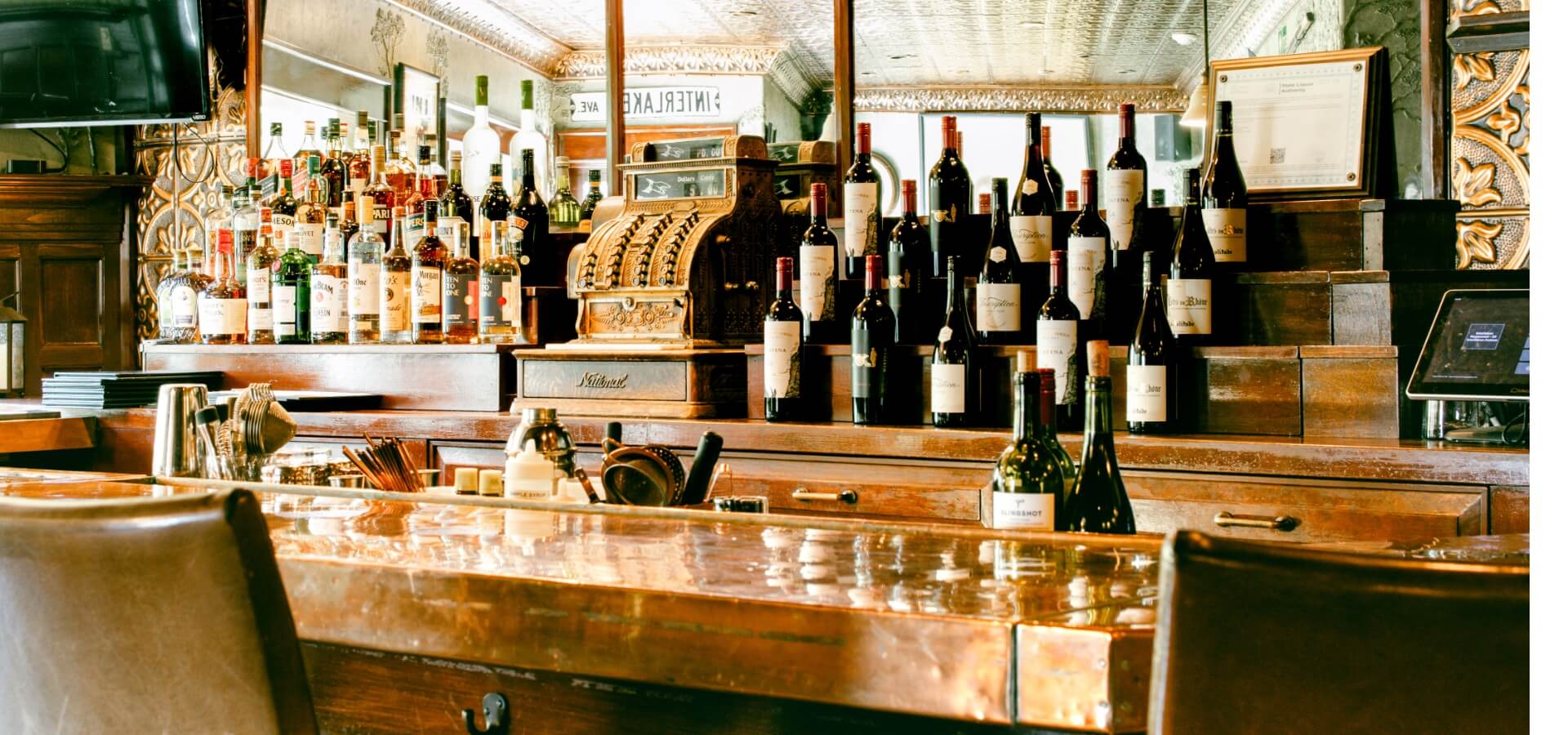 A wooden bar with an array of bottles and a vintage cash register in a warmly lit setting.