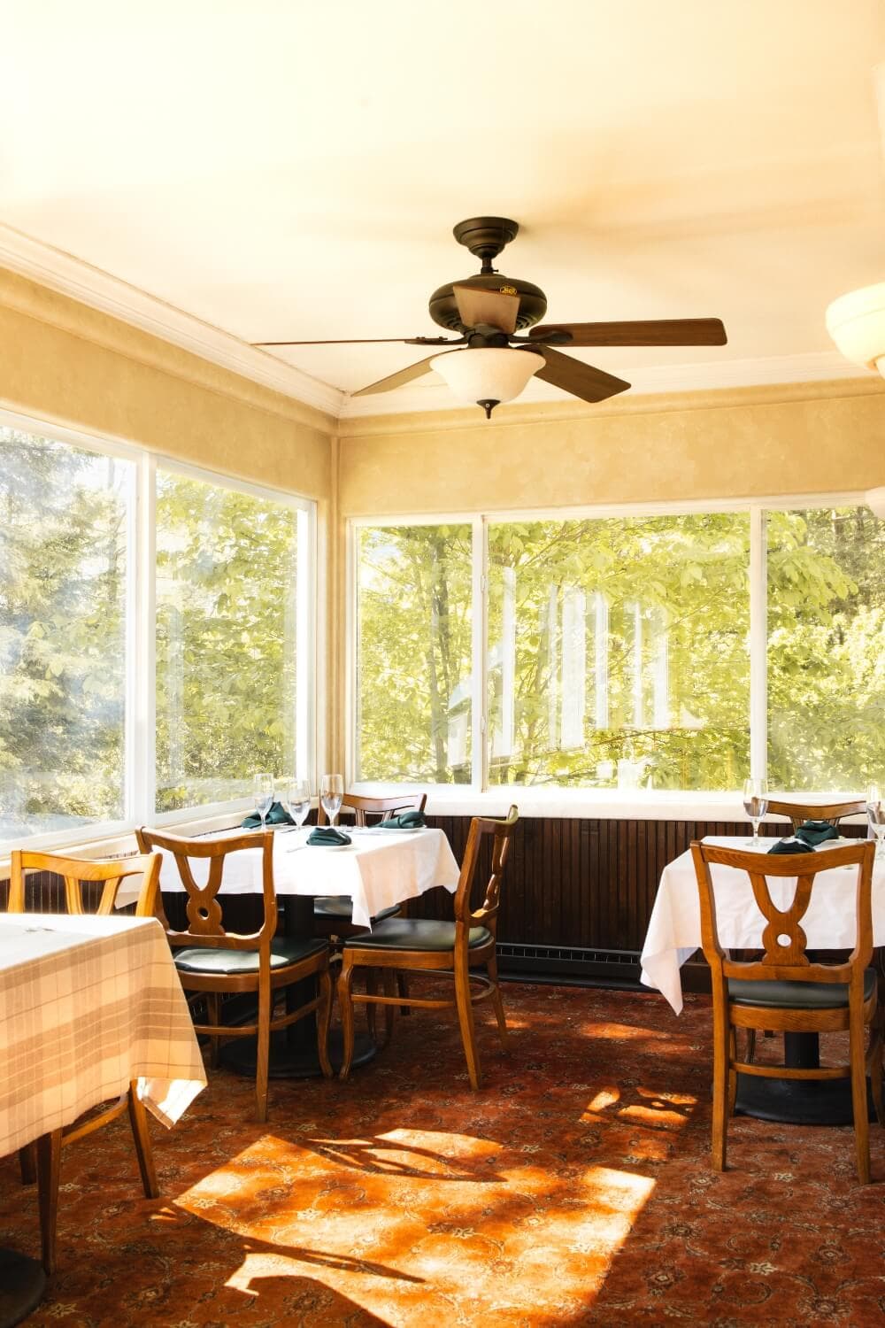 Bright dining area with wooden chairs and tables, featuring large windows overlooking greenery.