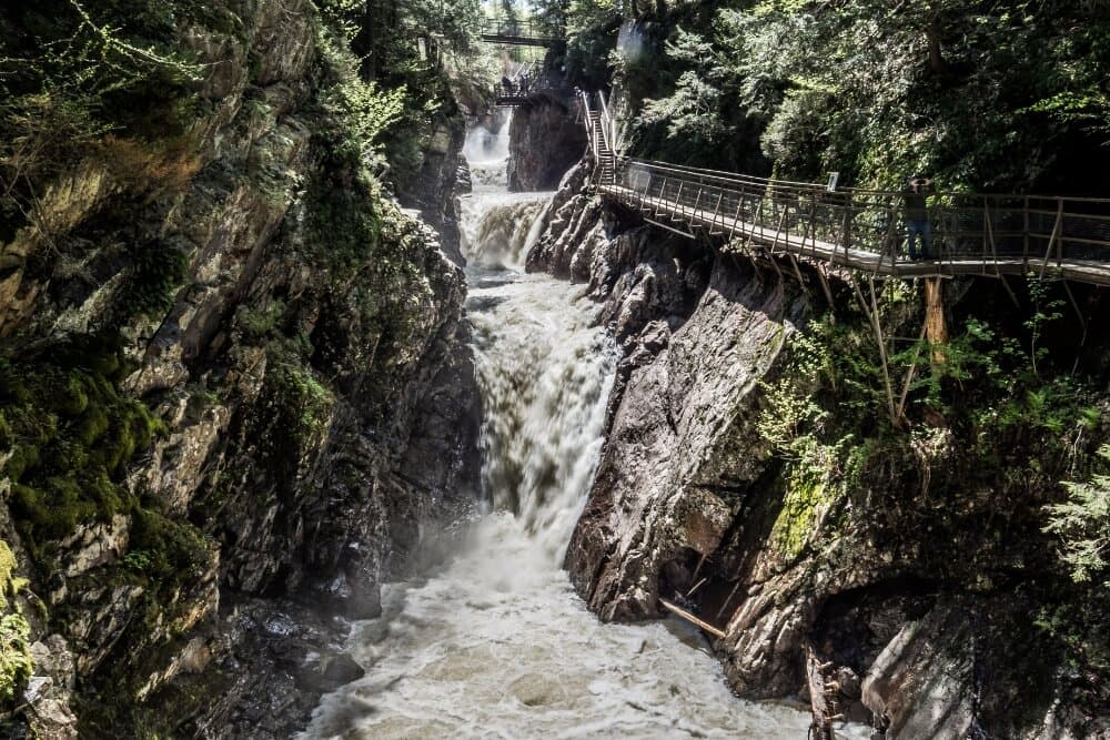 A waterfall cascades through a rocky gorge, with a wooden walkway above surrounded by lush trees.
