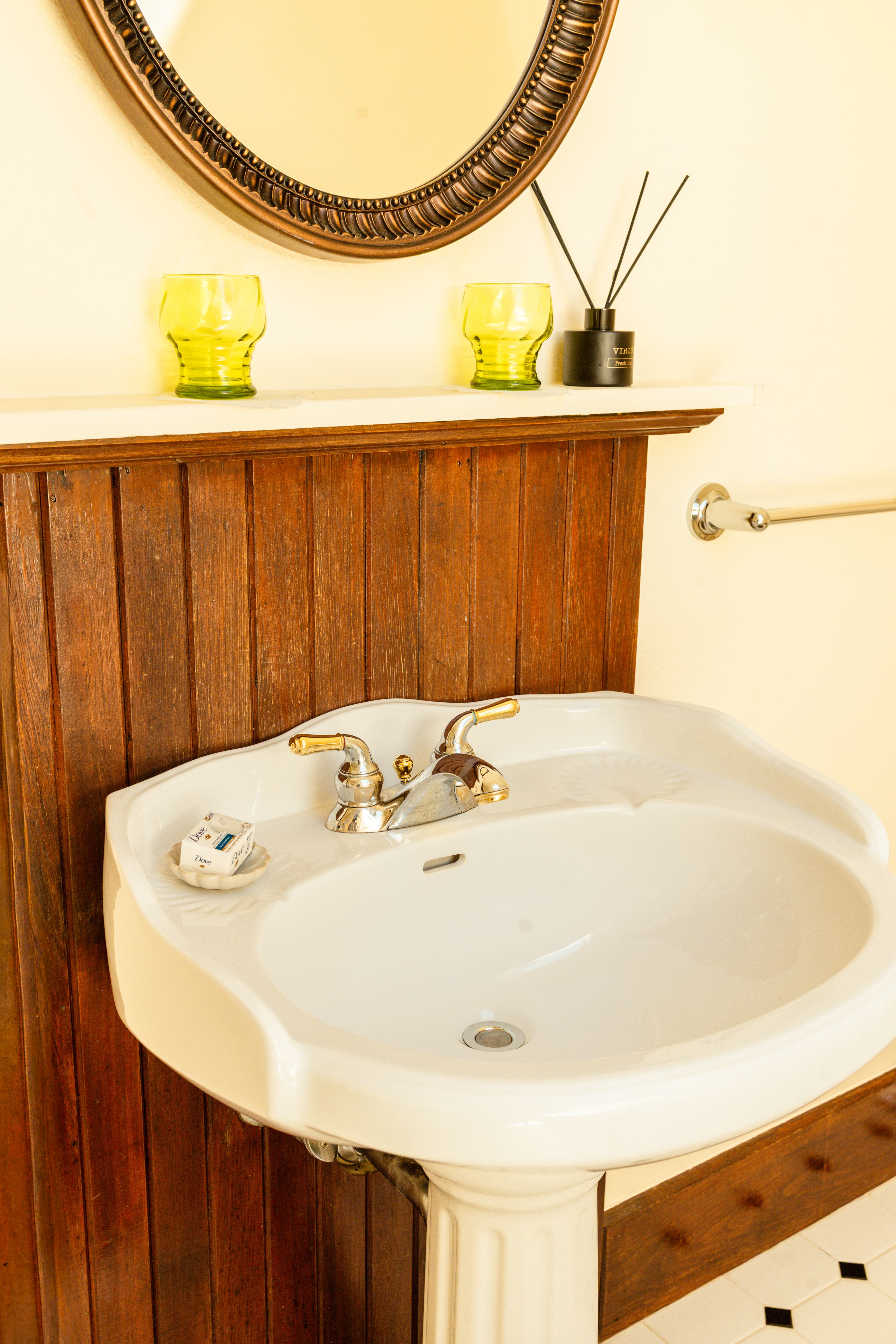 A white sink with gold faucets, next to yellow glass candles and a round mirror, set against a wooden wall.