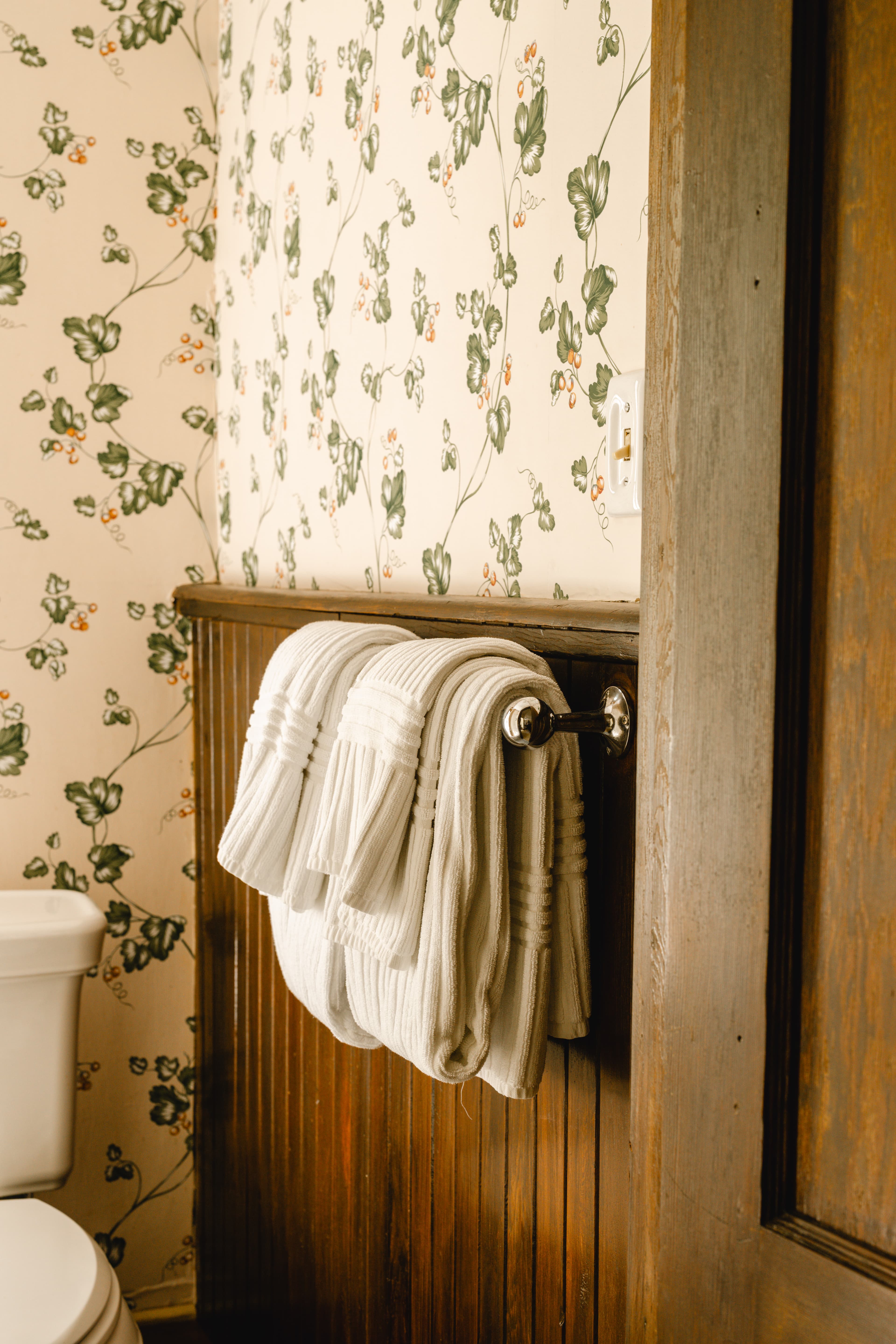 White towels hanging on a chrome rack in a bathroom with floral wallpaper.