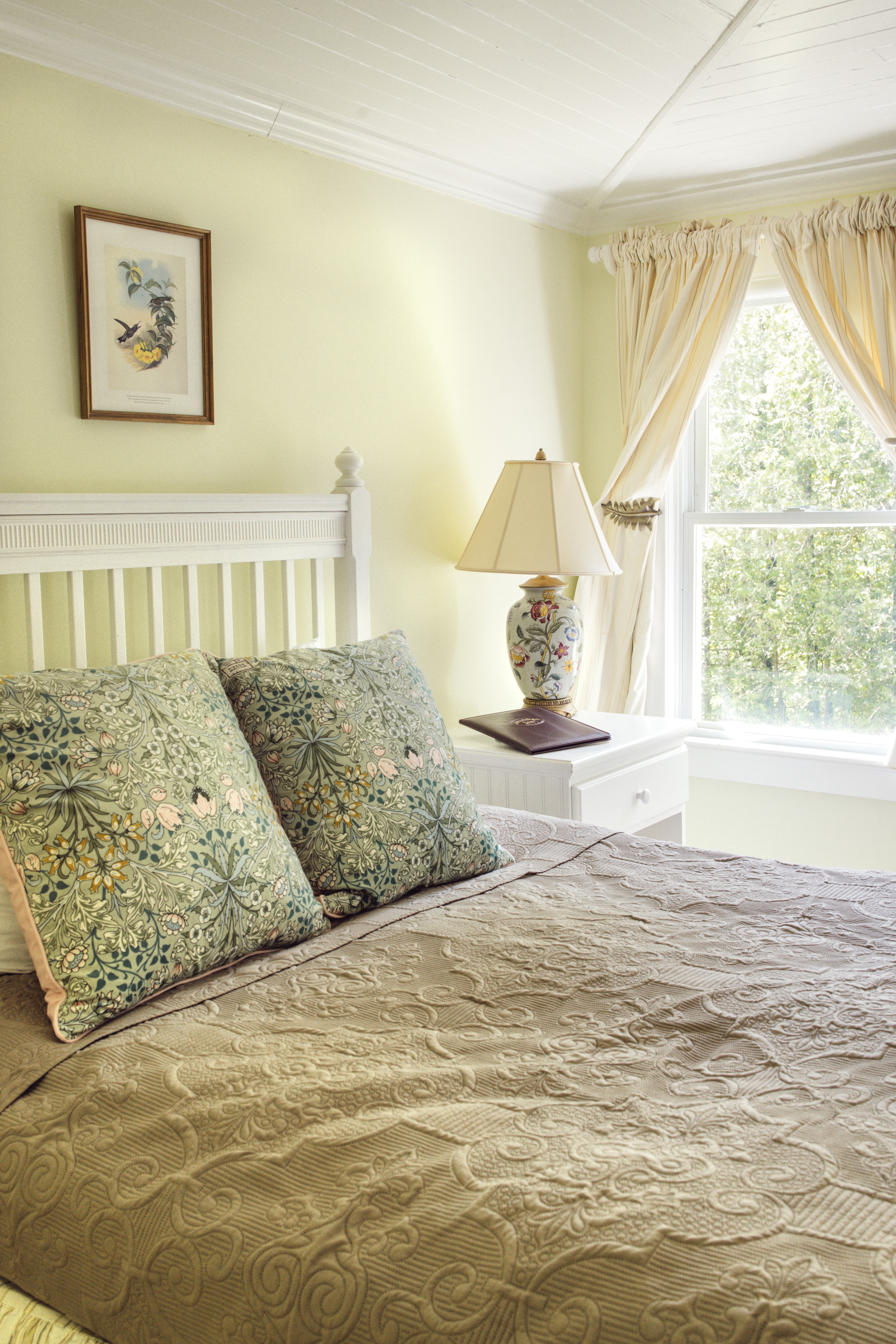 A cozy bedroom featuring a white bed with decorative pillows, a bedside lamp, and a window with soft curtains.